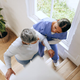 woman, nurse and helping senior up stairs in retirement home for support, trust or healthcare. female person, medical caregiver holding arms on staircase above with retired or mature patient at house